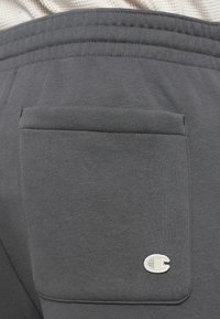 Tejido de sudadera gris con un detalle de bolsillos traseros. Presenta un pequeño logotipo bordado en un diseño circular en el bolsillo.