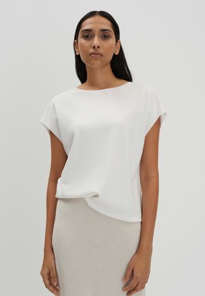 Witte top met korte mouwen en ronde hals, met een gedrapeerd ontwerp en een gladde textuur. Gedragen over een lichtbeige rok, wat een minimalistische look biedt.