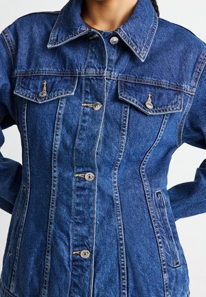 Gros plan d'une personne portant une veste en jean bleue cintrée avec des boutons métalliques et des poches à rabat sur la poitrine.