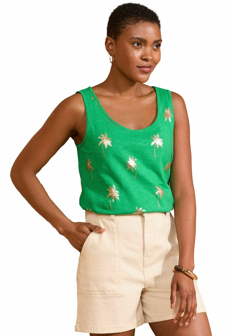 Top verde con ricami di palme dorate, scollatura a barca e tessuto morbido, abbinato a pantaloni beige chiaro e un braccialetto di legno.