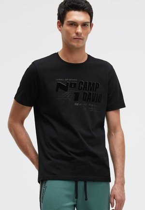 Mann trägt schwarzes Grafik-T-Shirt mit dem Text "No 1 Camp David" und türkisfarbene Hose mit Kordelzug, steht vor einfachem hellem Hintergrund.