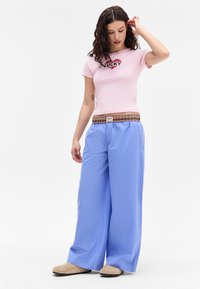 Rosa tettskjorte med "LUCKY" grafikk, parret med vide bukser med blå striper og brun rutet livlinje, komplettert med beige slip-on sko.