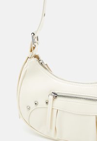 Krémová kabelka přes rameno z umělé kůže zaobleného tvaru, se zipovými kapsami, skládáním a stříbrnými kovovými detaily.