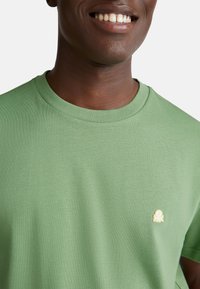 Usmívající se osoba v lehce zeleném tričku s malým žlutým vyšitým logem na hrudi před jednobarevným pozadím.