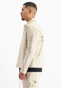 Beige cordjack met een gestructureerde afwerking, drukknoopsluiting en borstzak, gedragen over een zwart T-shirt en lichtbeige broek.