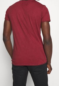 Camiseta de algodón burdeos de manga corta, con cuello redondo y un detalle rectangular cosido en la parte superior de la espalda.