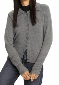 Šedý kardigan z měkké pletené látky. Má límec, zapínání na knoflíky a žebrované manžety. Nosit přes černý top.