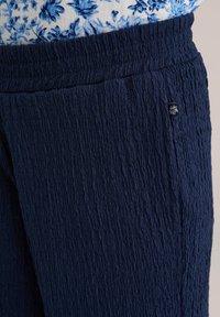 Navy textured pants with a crinkled surface, elastic waistband, and small embroidered logo on the side. Suitable for casual wear.