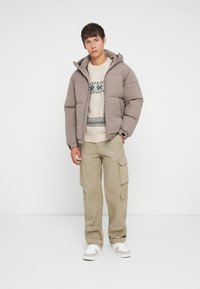 Tânăr îmbrăcat cu o geacă puffer taupe, un pulover cu model, pantaloni cargo bej și adidași albi, stând în fața unui fundal alb simplu.
