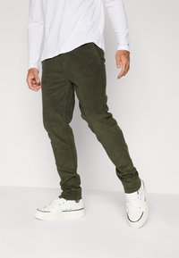 Olivgrön kordfl varpbyxor med avsmalnande ben, med framfickor, bärs med vita sneakers och en långärmad vit skjorta.
