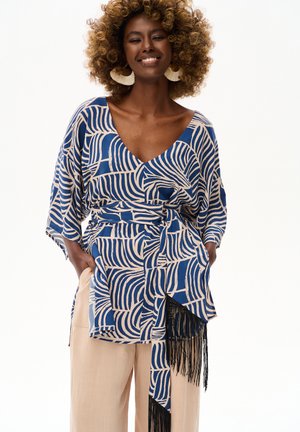 Uśmiechnięta kobieta, ubrana w niebiesko-beżową, geometrycznie wzorzystą bluzkę typu wrap z czarnymi frędzlami i beżowe szerokie spodnie, ręce w kieszeniach.