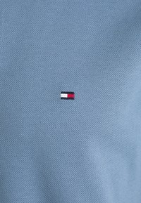 Blå texturerad tygpolo med en liten broderad logotyp med röd, vit och marinblå rand på vänster sida.