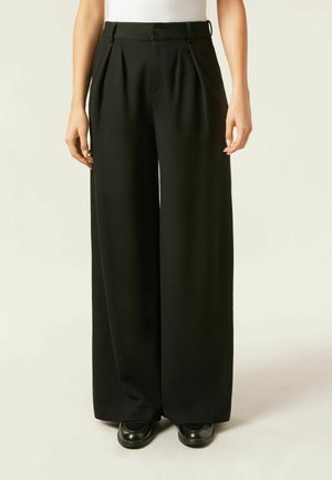 Pantalons larges noirs avec des plis, fabriqués à partir d'un tissu lisse. Présente une fermeture à bouton et des passants de ceinture, associés à des chaussures noires.