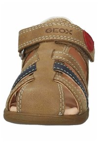 Sandale en cuir couleur tan avec design ouvert, bride ajustable, rayures bleu marine et coutures blanches. Comprend des trous de ventilation pour la respirabilité.