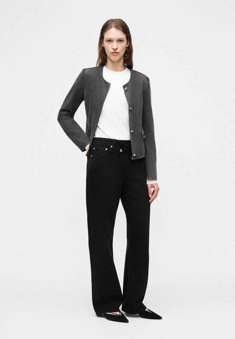 Femme portant une veste boutonnée gris foncé, une chemise blanche, un jean large noir et des ballerines noires à bout pointu, se tenant devant un fond blanc.