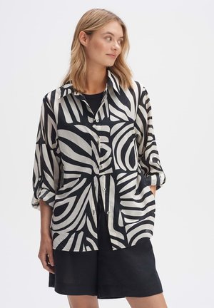 Camicia a righe bianco e nero con motivi astratti, realizzata in tessuto leggero, con abbottonatura frontale e maniche arrotolate.