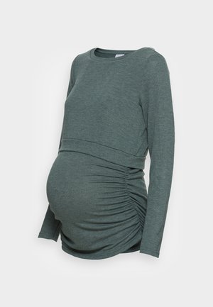 Tmavě zelený těhotenský top s dlouhým rukávem a nařasenými boky, navržený tak, aby padl přes rostoucí břicho.