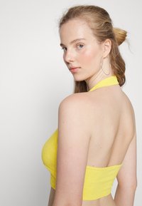 Top halter amarelo, texturizado com um design costas nuas. A modelo tem cabelo comprido e usa grandes brincos de argola prateados.