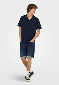 Mornarska polo majica s džepom, uparena s tamnim trapericama s obrezanim rubovima. Model nosi bijele čarape i mornarske tenisice.