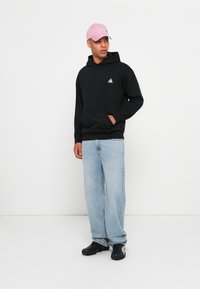 Schwarzer Hoodie mit einer Fronttasche und einem kleinen dreieckigen Logo, kombiniert mit hellblauen weiten Jeans und schwarzen Schuhen. Eine rosa Kappe rundet den Look ab.