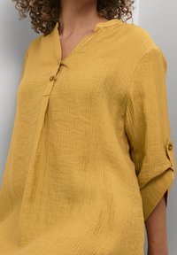 Senfgelbe, strukturierte Bluse mit V-Ausschnitt, Knopfverschluss und hochgerollten Ärmeln mit Knopfakzenten.