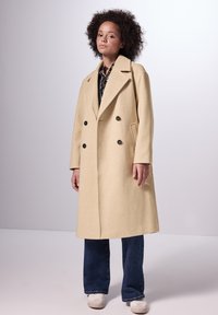 Cappotto di lana beige a doppio petto con ampi revers, grandi bottoni neri, tasche laterali, che arriva fino a metà polpaccio, abbinato a jeans scuri e sneakers bianche.