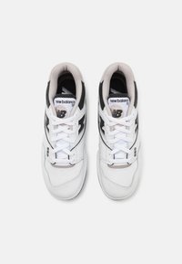 Fehér és fekete tornacipők bőr felsőrésszel, fűzős záródással, perforált orral és fémes részletekkel. Az "new balance" logó és a modell szám is látható.