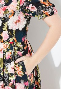 Die Hand einer Frau ruht in der Tasche eines schwarzen Kleides mit pinken, orangen und grünen Blumenmustern und puffierten kurzen Ärmeln.