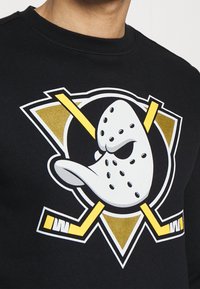 Svart sweatshirt med en stor grafik av en hockeymask med gula och vita accenter, mot en triangel-logotypbakgrund.