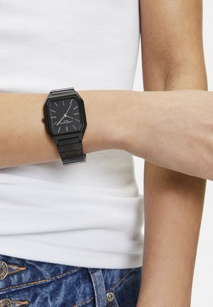 Schwarze rechteckige Armbanduhr mit Metallarmband, getragen am linken Arm über einem weißen Tanktop und blauen Jeans.