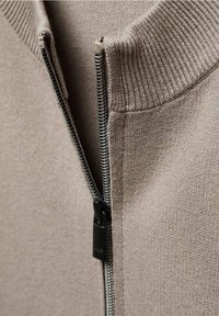 Beige zip-up sweater featuring a ribbed collar, smooth texture, and metal zipper with a pull tab, providing a close-up view of the fabric.