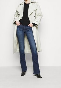 Manteau trench gris clair avec un large col, pull en laine noir, jean évasé bleu moyen et chaussures à talons noires.
