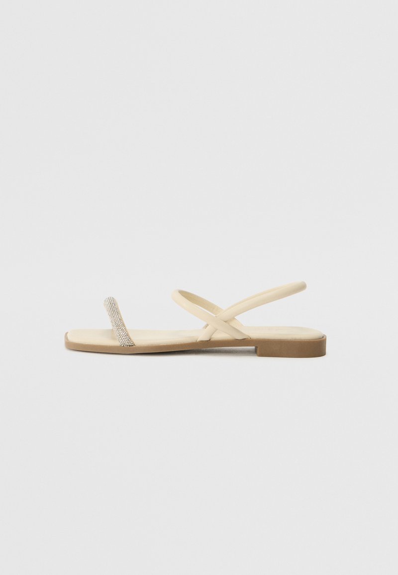 Sandalettes beiges à semelles plates avec des lanières croisées et un anneau pour les orteils orné de strass. Dispose d'une semelle lisse et d'un design minimaliste.