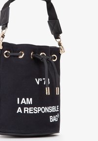 Le sac seau en toile noire présente un texte blanc, des accessoires en or, un fermeture à cordon, et une sangle texturée avec des accents matelassés. Ajustable et compact.