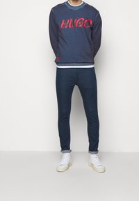 Felpa navy con grafica rossa "HUGO", collo a coste e polsini; abbinata a jeans skinny in denim scuro e sneakers bianche.