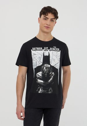 Maglietta nera in cotone con grafica "BATMAN VS THE JOKER". Il design include illustrazioni audaci di Batman e Joker in scala di grigi.
