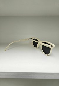 Zonnebril met een crèmekleurig plastic frame, zwarte glazen en een ronde vorm. Heeft een slanke poot met merkdetails.