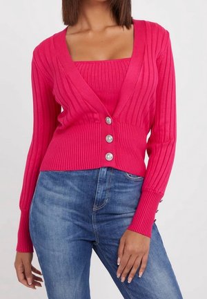 Femme porte un cardigan côtelé rose vif avec trois boutons sur un haut sans manches assorti, associé à un jean denim bleu sur fond blanc.