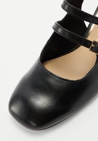 Chaussure en similicuir noir avec un bout rond, dotée de deux fines lanières et d'une boucle. L'intérieur est de couleur beige clair avec une texture lisse.