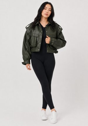 Femme aux longs cheveux foncés portant une veste courte en cuir vert foncé, leggings noirs et baskets blanches, debout devant un fond uni.