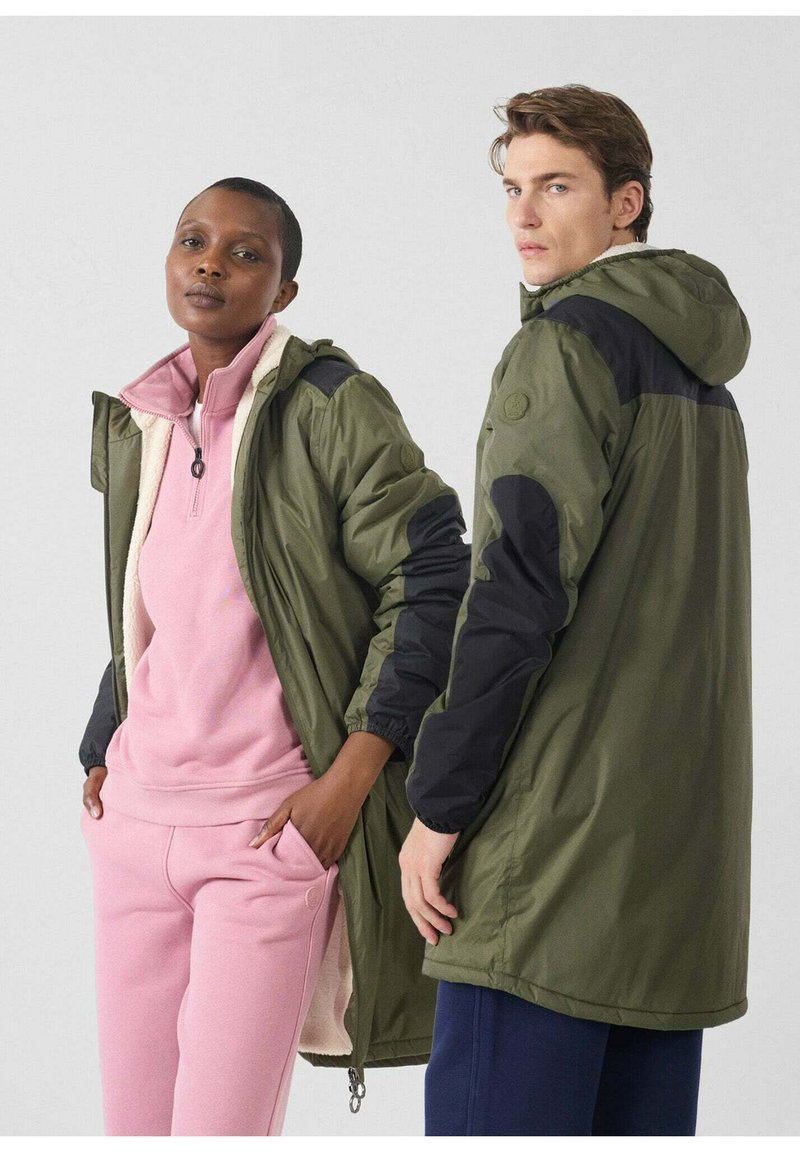 Olijfgroene jassen met zwarte accenten; de voorkant heeft een ritssluiting, terwijl een roze fleece top en bijpassende broek de look compleet maken.