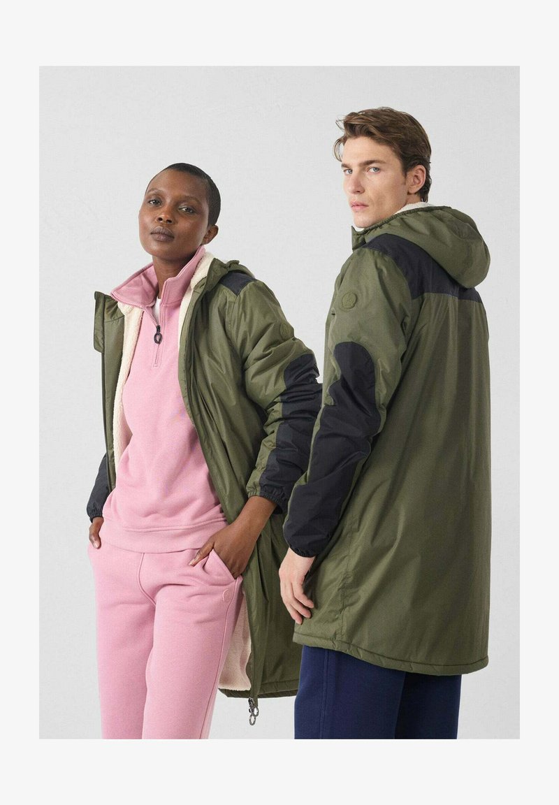 Olijfgroene jassen met zwarte accenten; de voorkant heeft een ritssluiting, terwijl een roze fleece top en bijpassende broek de look compleet maken.