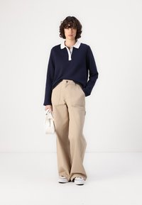 Marineblå langermet polo med hvit krage, kombinert med beige vide bukser. Modellen holder en liten hvit veske og står foran en nøytral bakgrunn.