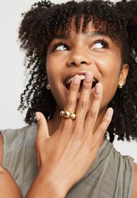Joyería en tono dorado que incluye un anillo esférico y pendientes a juego. El anillo tiene múltiples detalles esféricos que reflejan la luz. Cabello rizado con un estilo natural.