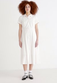 Rochie midi albă cu croi petrecut și mâneci scurte, realizată dintr-un material ușor, având un tors ajustat și fustă evazată, completată cu pantofi negri și șosete albe.