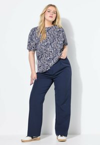 Giovane donna in posa con una maglietta a maniche corte blu con motivo paisley, pantaloni larghi blu scuro e scarpe da ginnastica bianche su uno sfondo bianco.