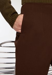 Brązowe spodnie mają gładką fakturę i boczną kieszeń, a do nich dołączony jest oliwkowy sweter z prążkowanym wzorem.