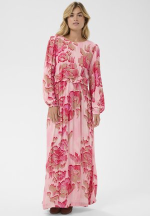 Kobieta stojąca, ubrana w długą sukienkę w różowe i czerwone kwiaty, z długimi rękawami i falbanką w talii, na białym tle.