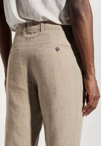 Bežové lněné kalhoty s volnějším střihem, které mají dvě zadní kapsy se zapínáním na knoflíky a pás s poutky na opasek. Viditelná strukturovaná tkanina.