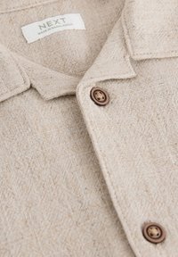 Chemise en lin beige à surface texturée, avec un col, des boutons marron et une étiquette indiquant "NEXT, FABRIQUÉ AU BANGLADESH".